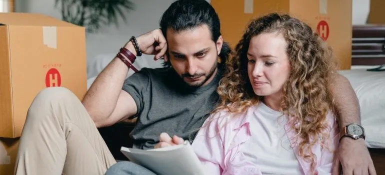 A couple sitting and writing down things in a notebook with boxes for the move behind them.