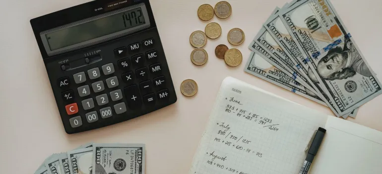 a black calculator next to a stash of dollars and a notepad of expense calculations