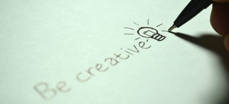 a person writing "be creative" on paper with a pencil